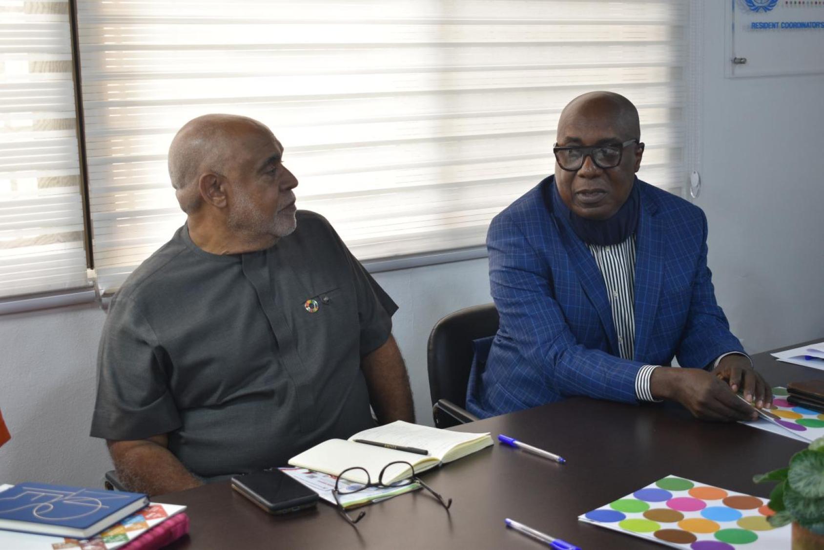 A man in a grey shirt and an SDGs pin talks to another man in a blue suit.