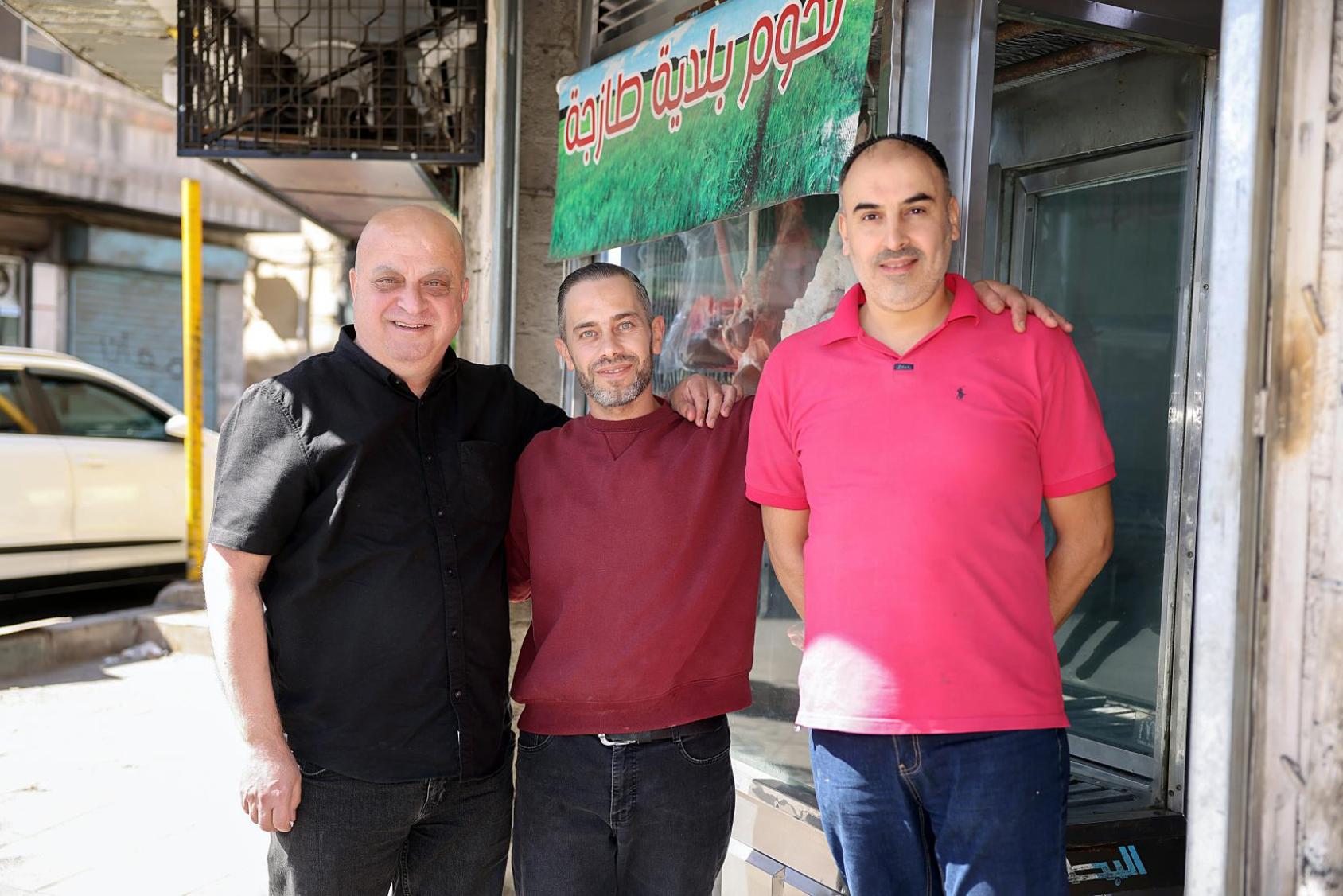 Three men, one in a red shirt, one in a black shirt and one in a pink shirt stand in front of a shop window