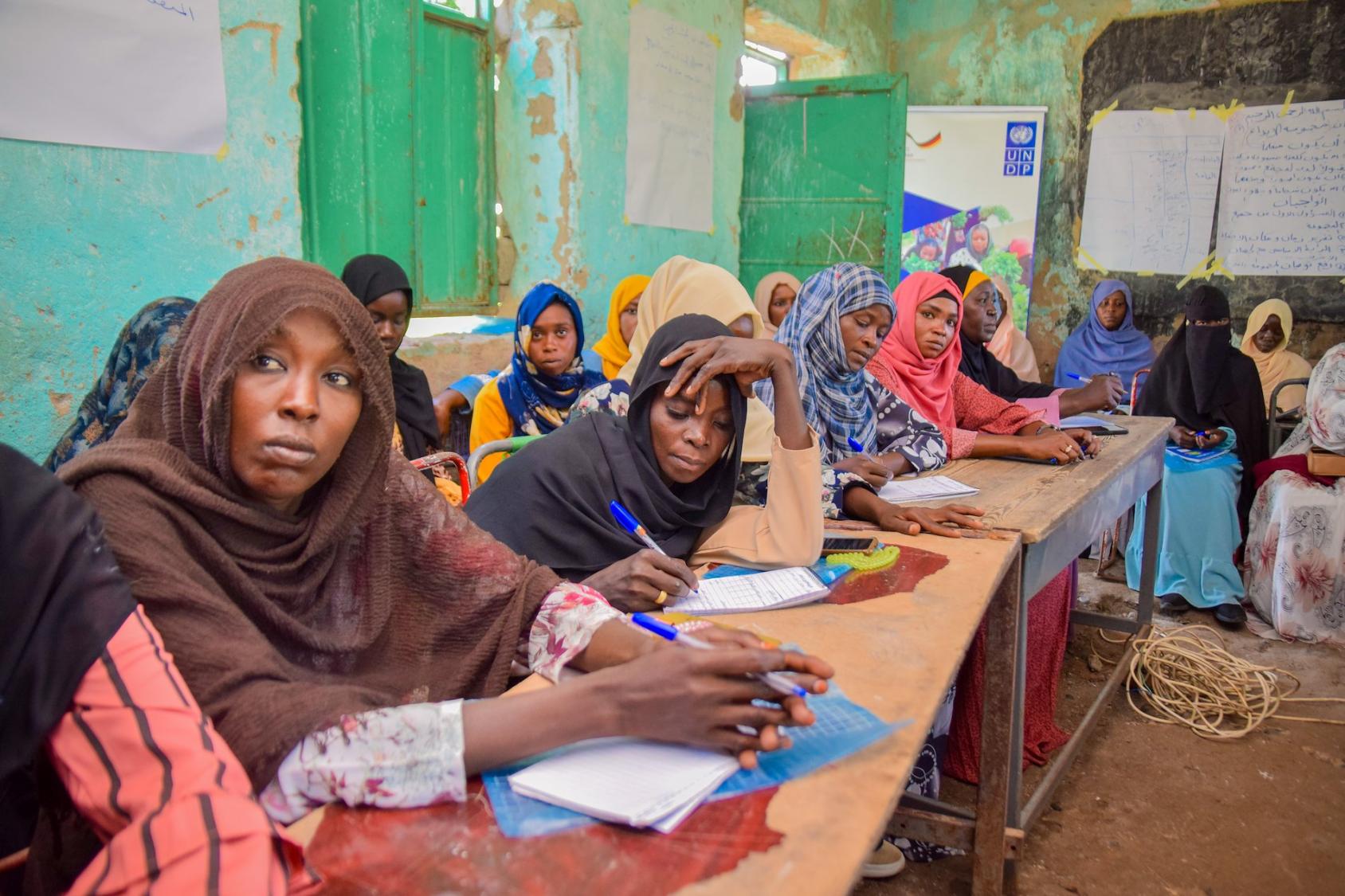 A classroom where several women in colourful clothes sit at desks next to each other, making notes in open notebooks.