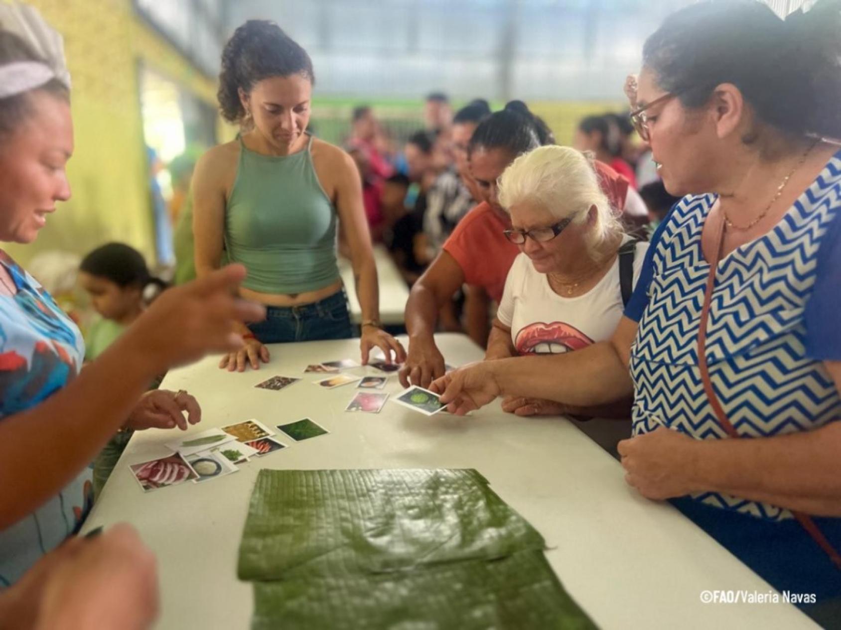 Four women gather around a table, reading cards and wrapping tamales in banana leaves