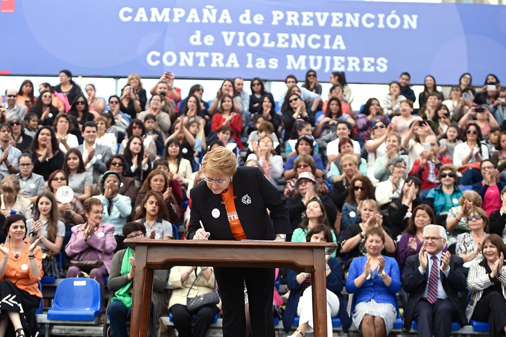 A woman in an orange shirt and black blazer stands at a podium and signs a document while a crowd of men and women behind her look on.