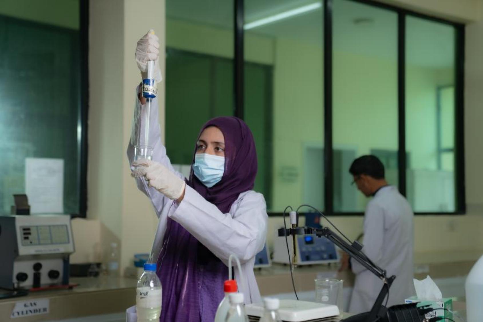 A woman scientist in a purple robe and headscarf, a lab coat and a mask and gloves uses a pipette to test water in a tube.
