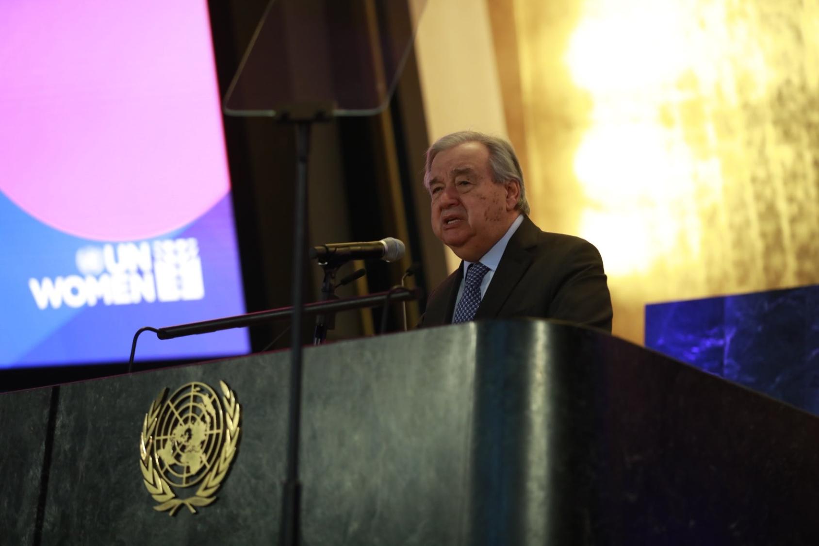 A man in a black suit and blue tie stands at a podium with the UN logo below it