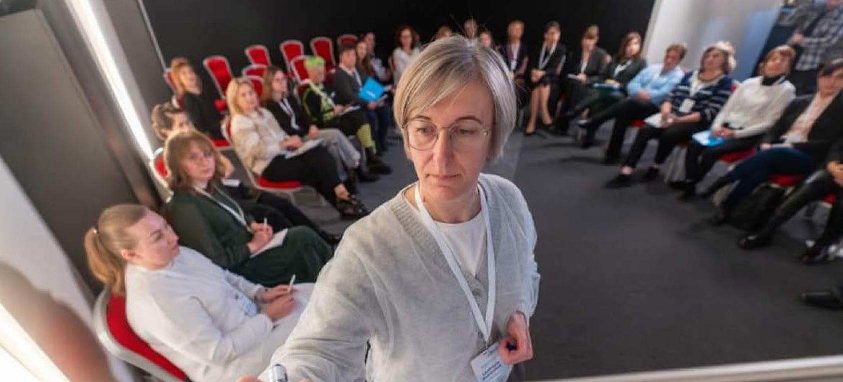 一位女士站在一个房间的前方似乎在书写什么，其他人环绕在其身后坐着，听讲中