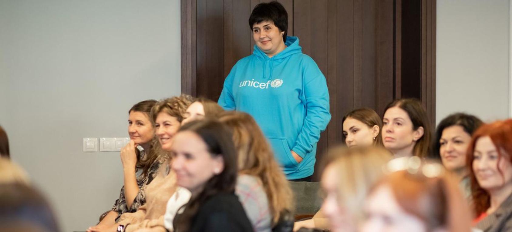 一位身穿联合国儿童基金会衣服的工作人员站在一个教室的后门，其他年轻人在教室里坐着，往向前方