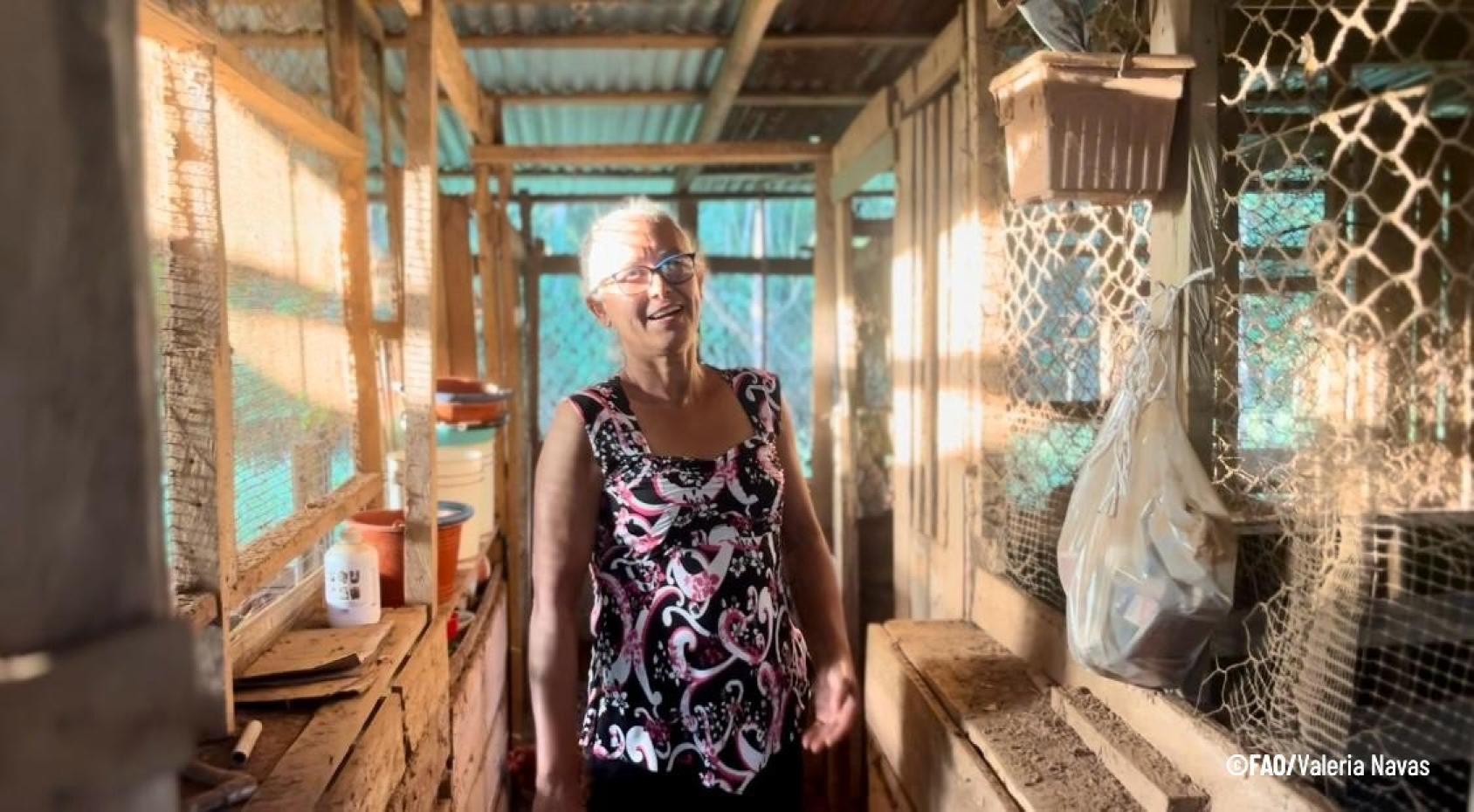 A woman with white hair and glasses and a printed shirt stands in a wooden house