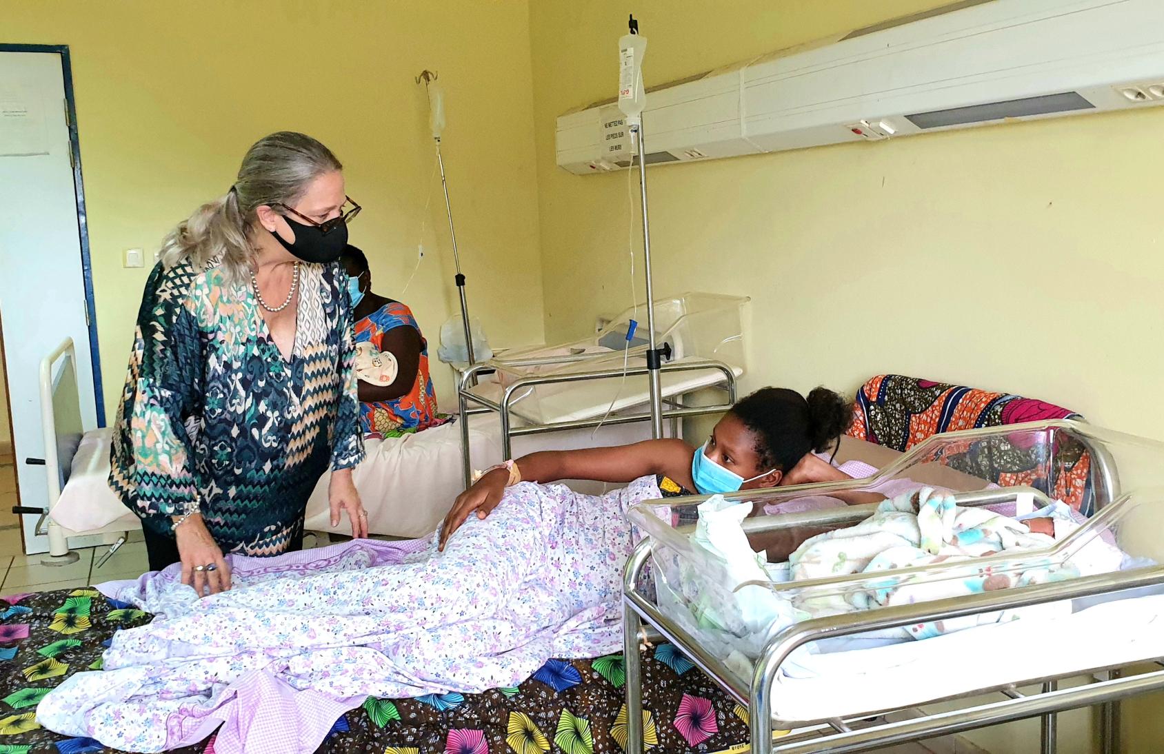 A woman in a printed dress and wearing a black face mask talks to another woman, a young mother, lying on a hospital bed with a child next to her.
