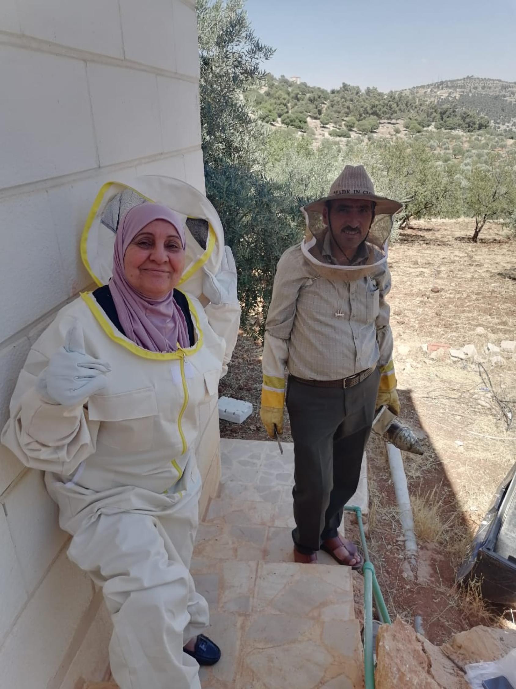 A woman in a beekeeping suit and a man in a grey uniform standing against a white wall.