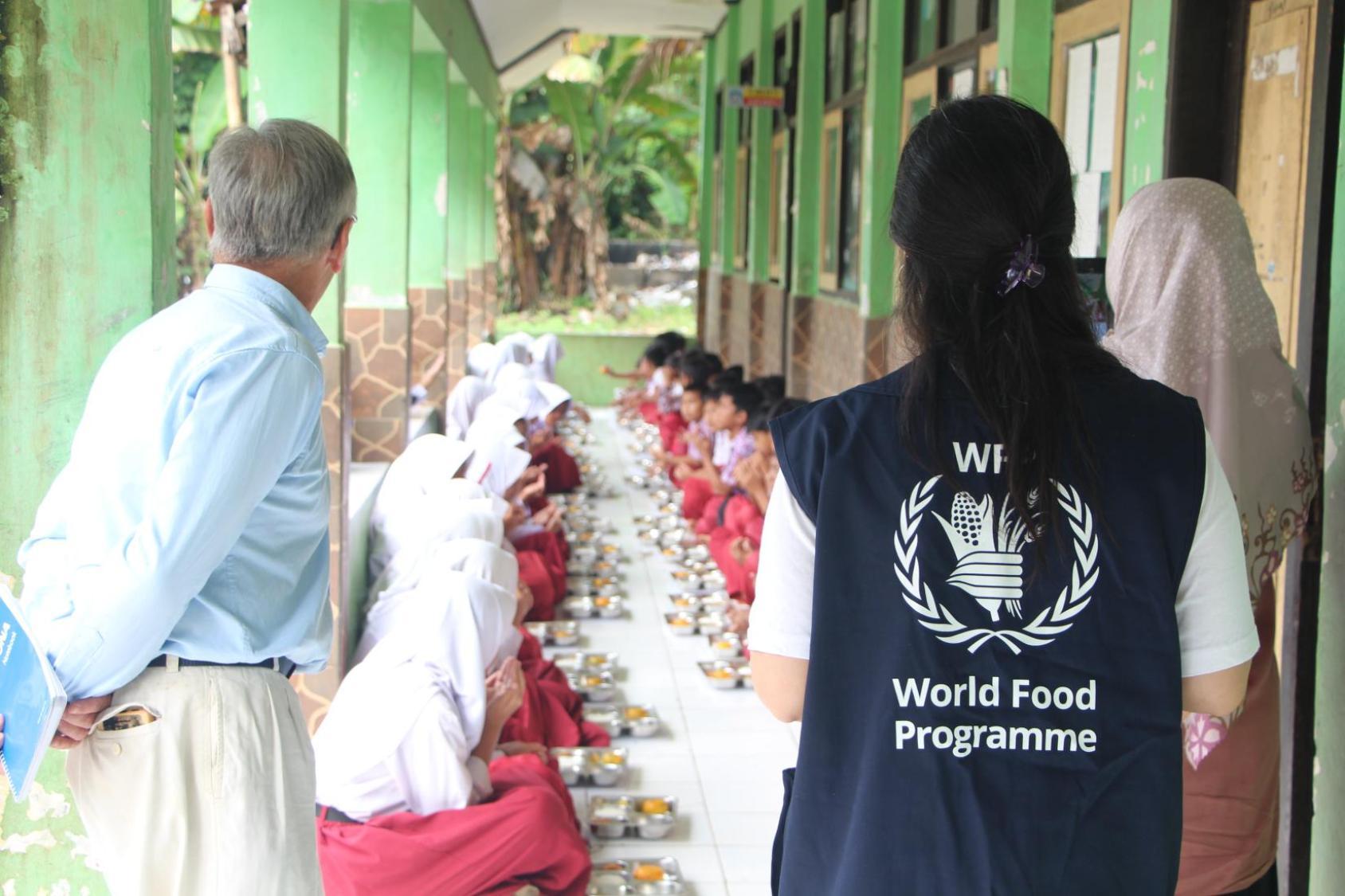A WFP staffer watches on as students eat on the floor
