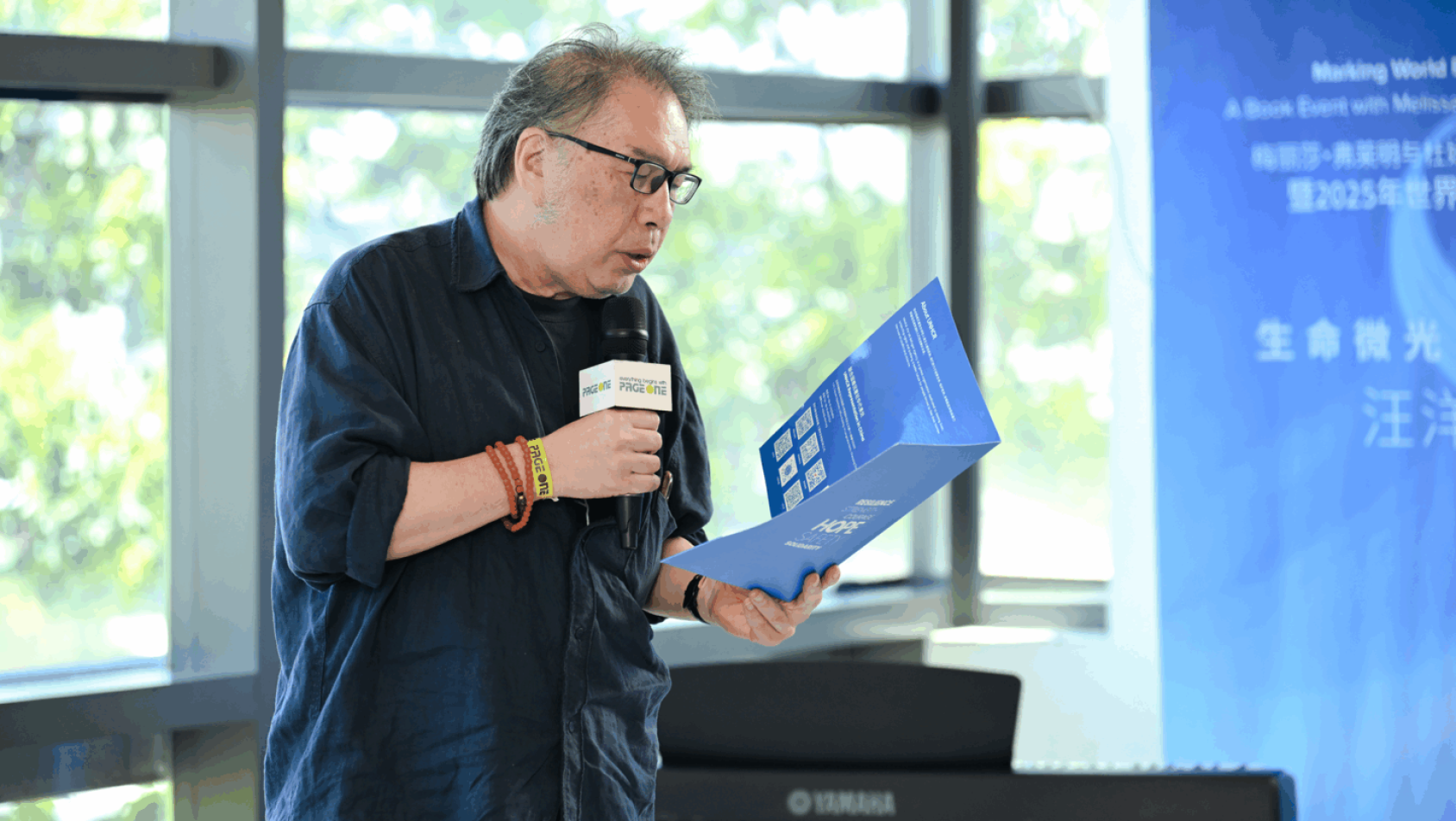 一位身着深色上衣戴着眼镜的男士手持话筒朗读一本书的内容