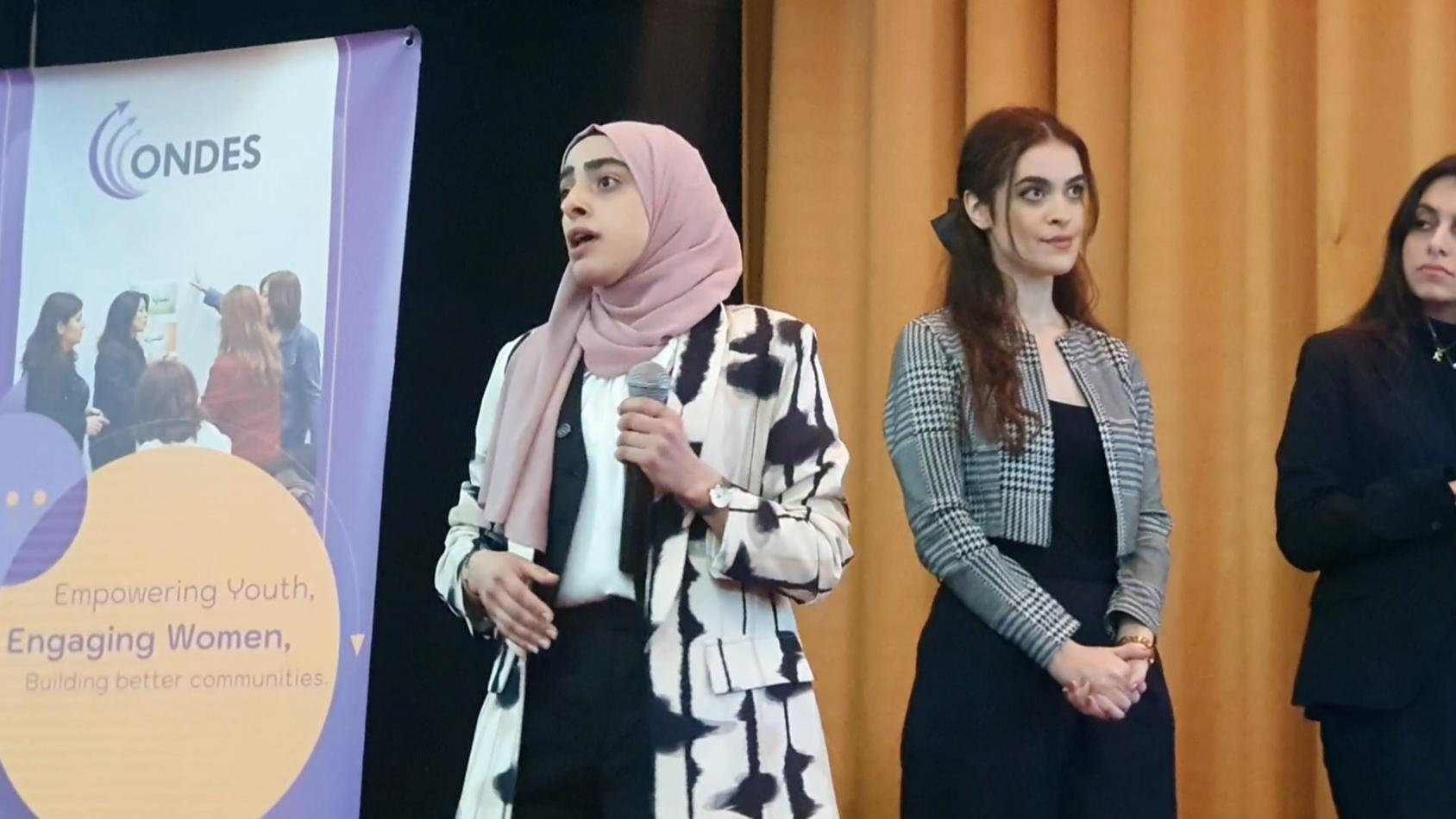 A woman in a pink headscarf in formal clothes talks to an audience while two other women standing behind her watch on.