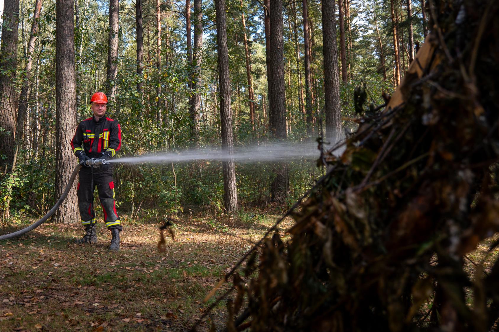 Лесник в защитном костюме и каске поливает лесную поляну водой из шланга