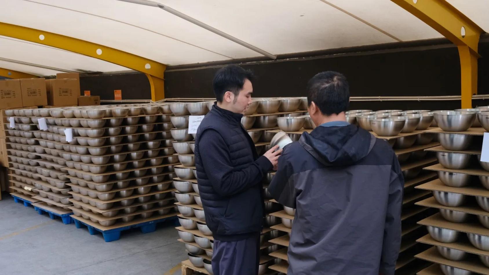 Dos trabajadores revisan y comentan recipientes metálicos apilados en estanterías dentro de un almacén o área de producción, en un contexto industrial y de control de calidad.