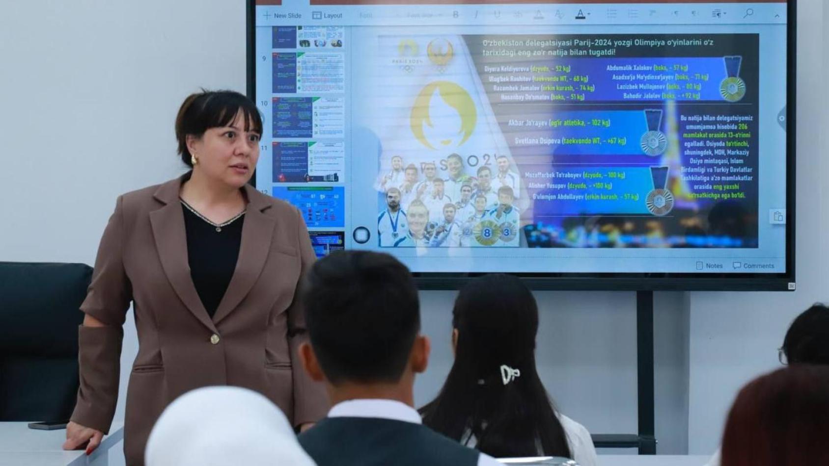 Женщина стоит у монитора с информацией и выступает с речью перед аудиторией.