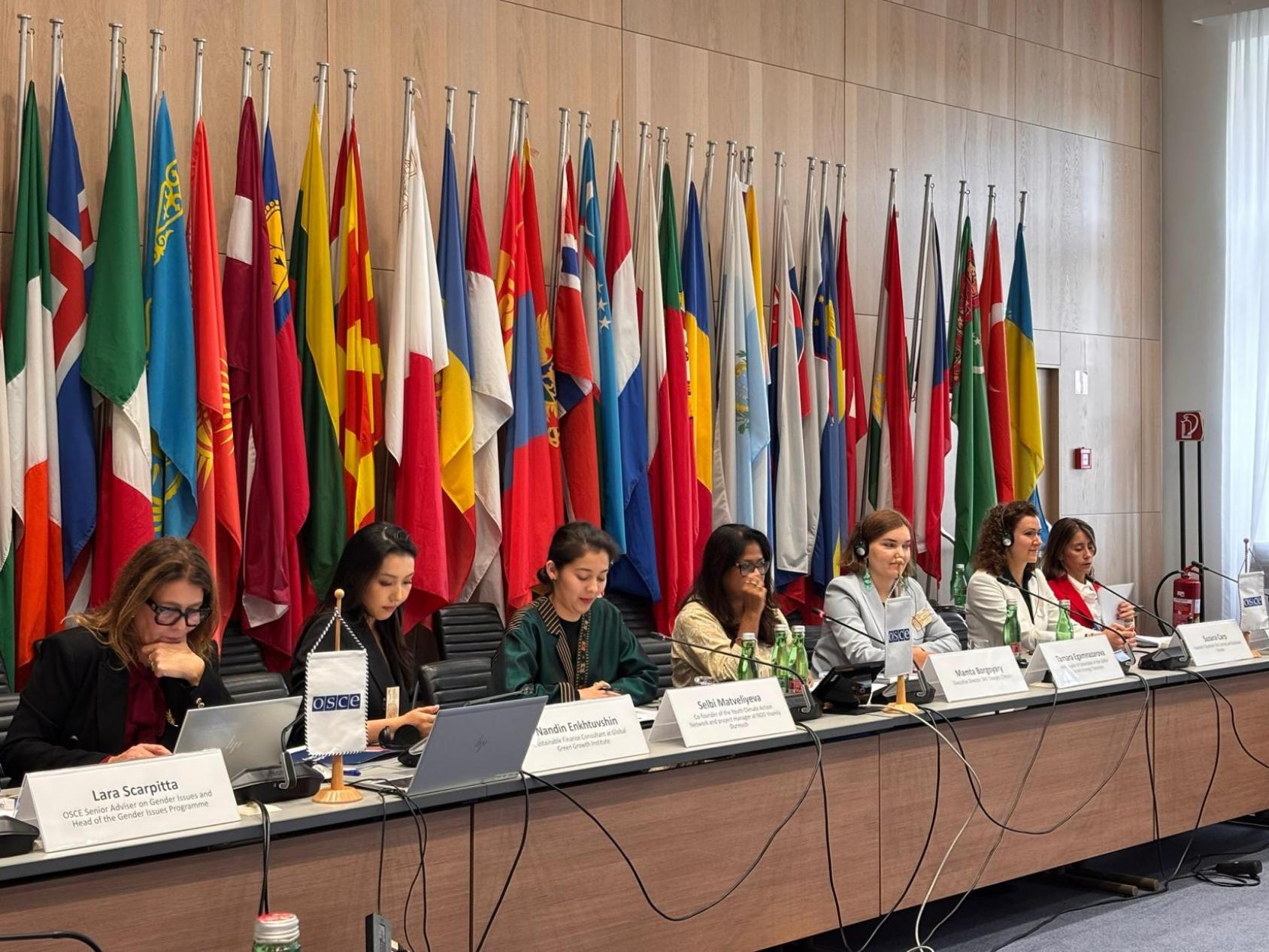 Девушки сидят за столом во время конференции на фоне флагов разных стран.