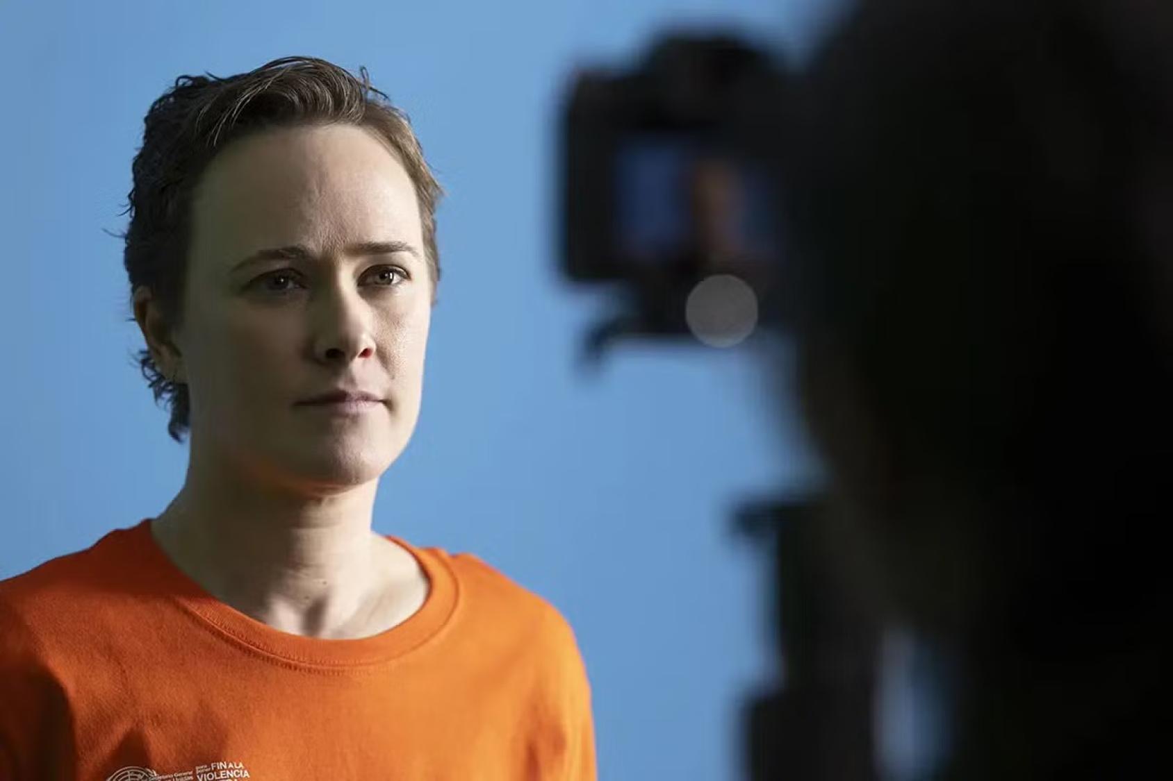 Una persona con camiseta naranja mira con expresión seria a una cámara que la graba en primer plano, sobre un fondo azul.