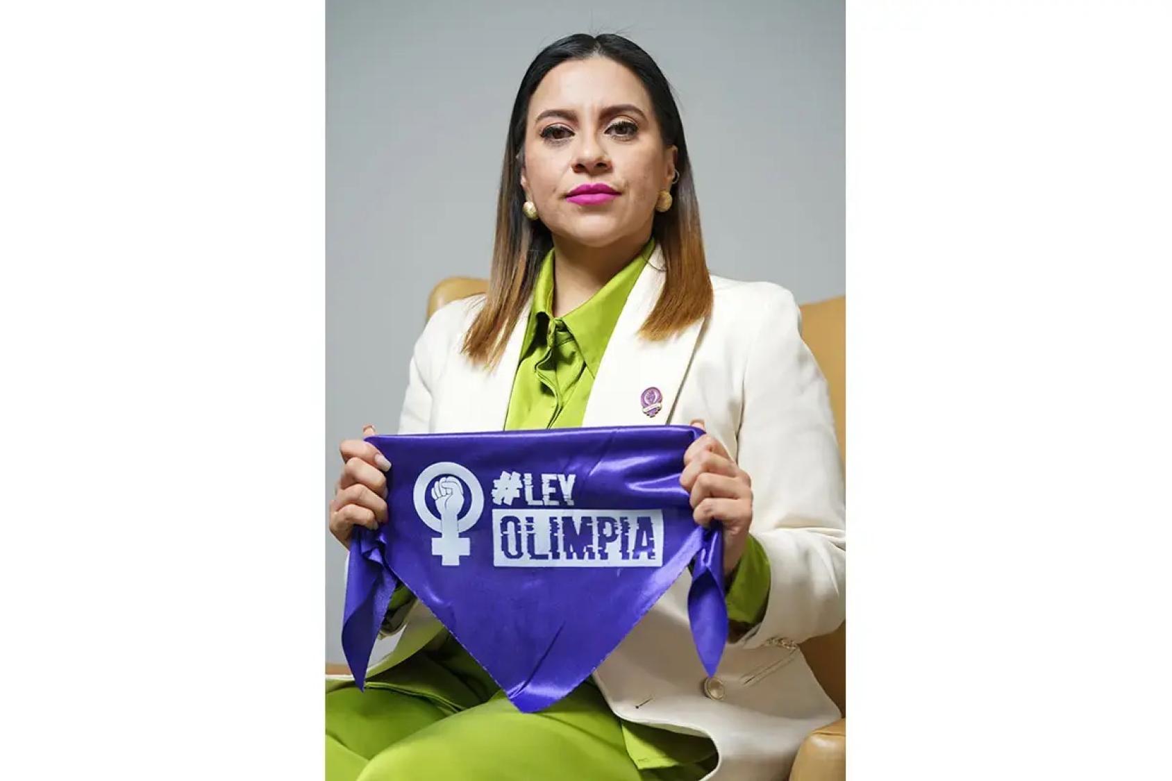 Una mujer vestida con traje claro y blusa verde sostiene frente a ella un pañuelo morado con el símbolo feminista y el texto “#Ley Olimpia”.