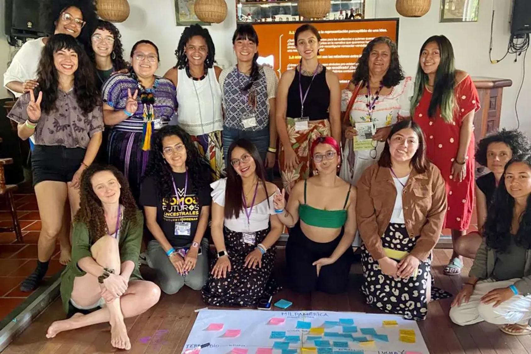 Un grupo numeroso de mujeres posa sonriente en un espacio interior tras una actividad, con notas de colores en el suelo y una pantalla con texto al fondo.