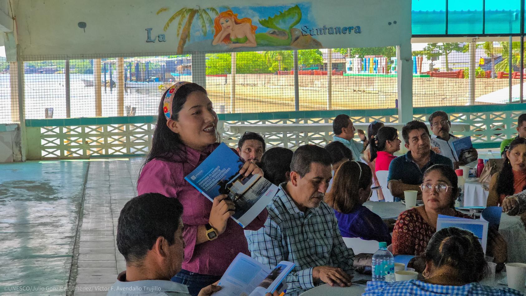 Una mujer de pie habla a un grupo de personas sentadas alrededor de mesas.