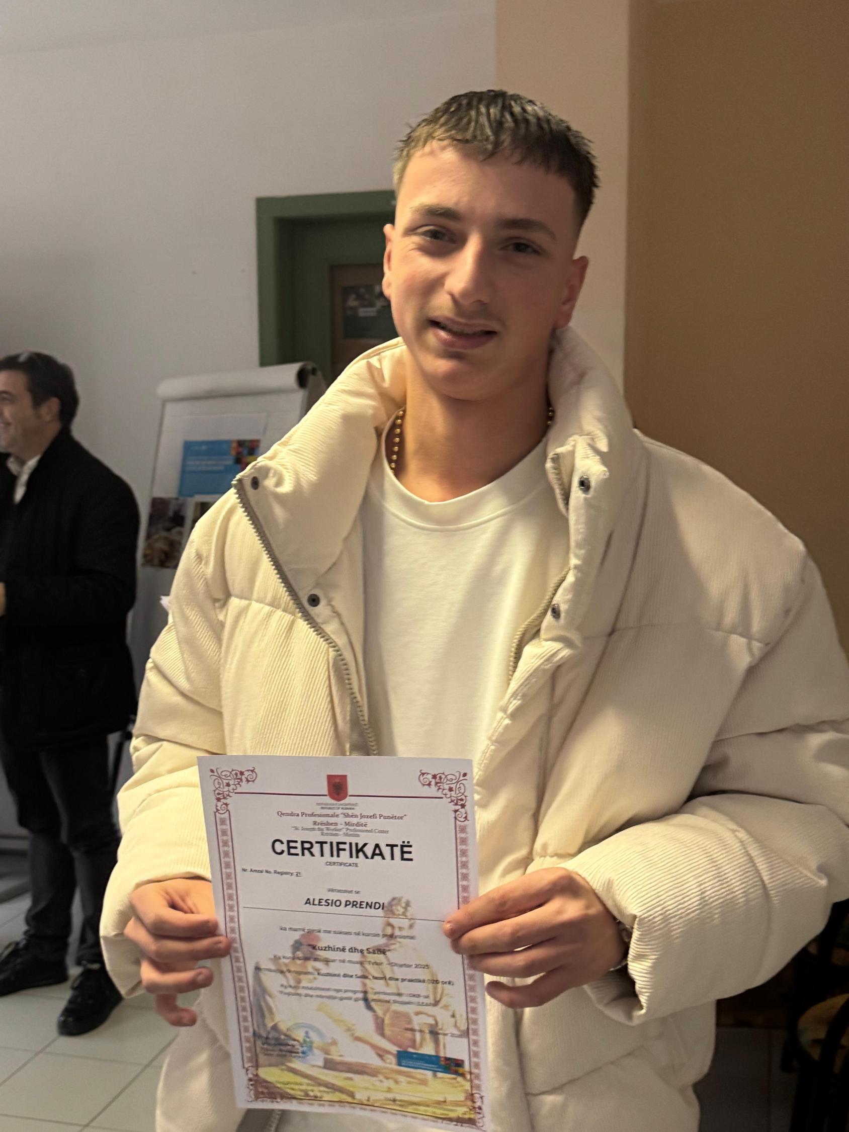 Молодой человек в белой куртке держит сертификат двумя руками и улыбается.