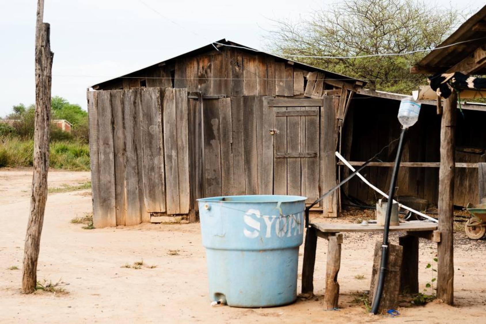 Una caseta de madera en un entorno seco, con un barril azul y una mesa rústica en primer plano.