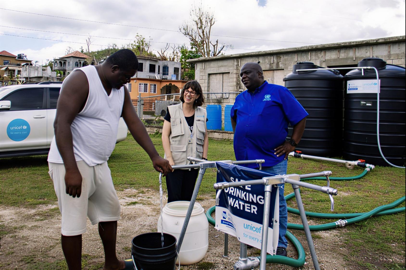 A man uses a water pump in front of two UN officials after a hurricane.