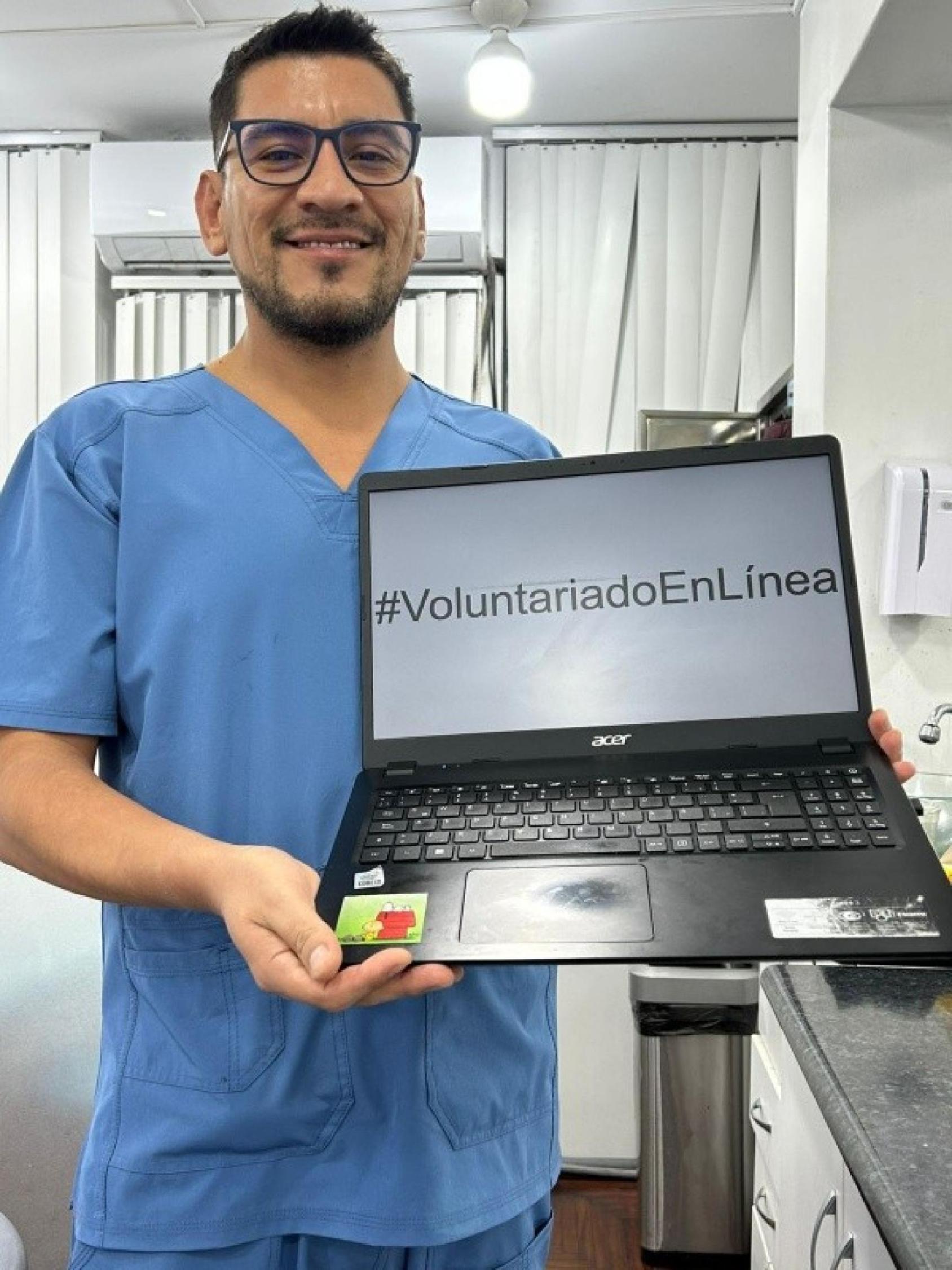 Un hombre con uniforme sanitario sonríe mientras sostiene una computadora en la que se lee “#VoluntariadoEnLínea”.