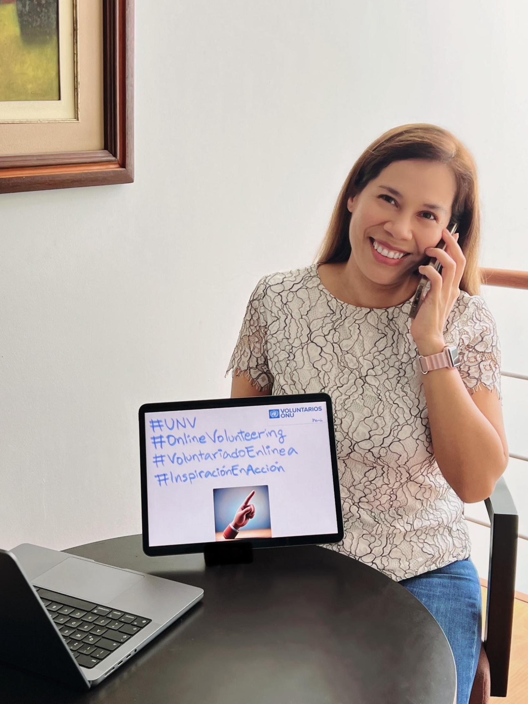 Una mujer sonríe mientras habla por celular junto a una tableta que muestra mensajes sobre voluntariado en línea.