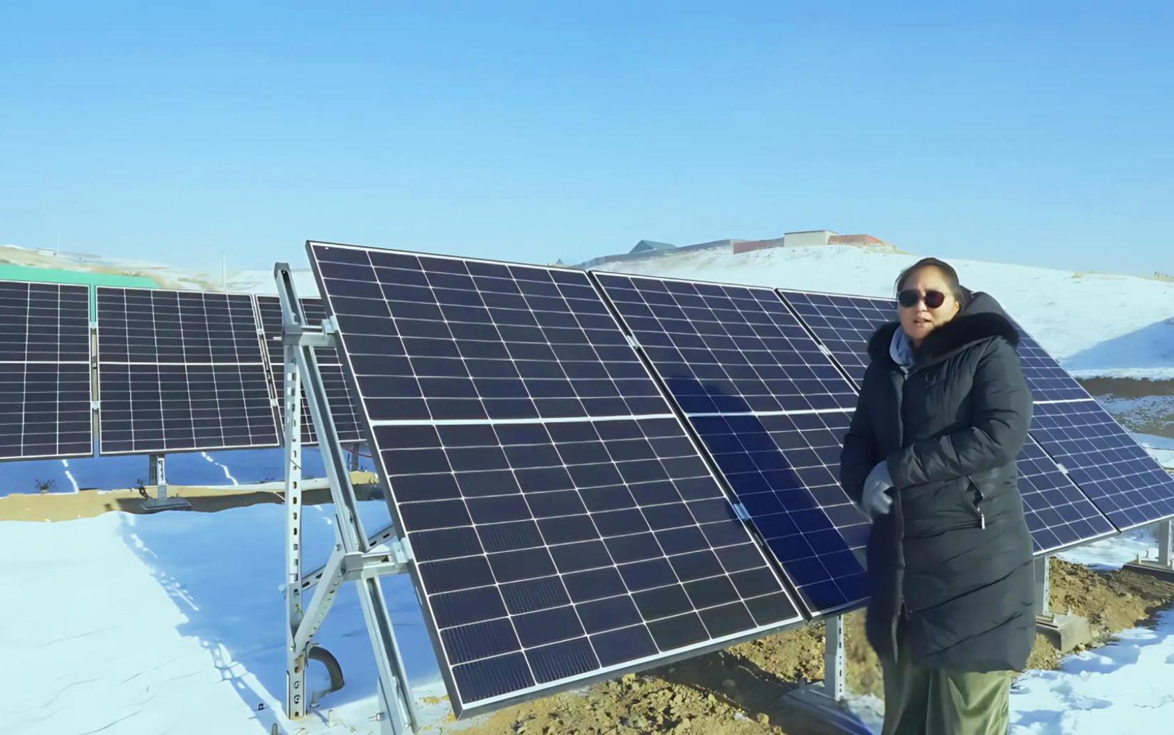 Женщина в зимней одежде, стоящая рядом с массивом солнечных панелей на заснеженном открытом пространстве.