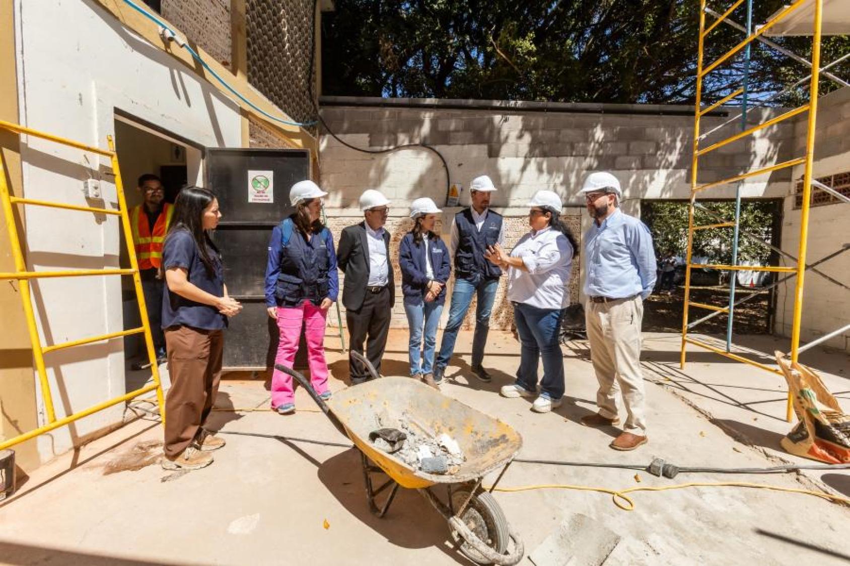 Un grupo de personas con cascos de obra conversa en un sitio en construcción, junto a una carretilla.