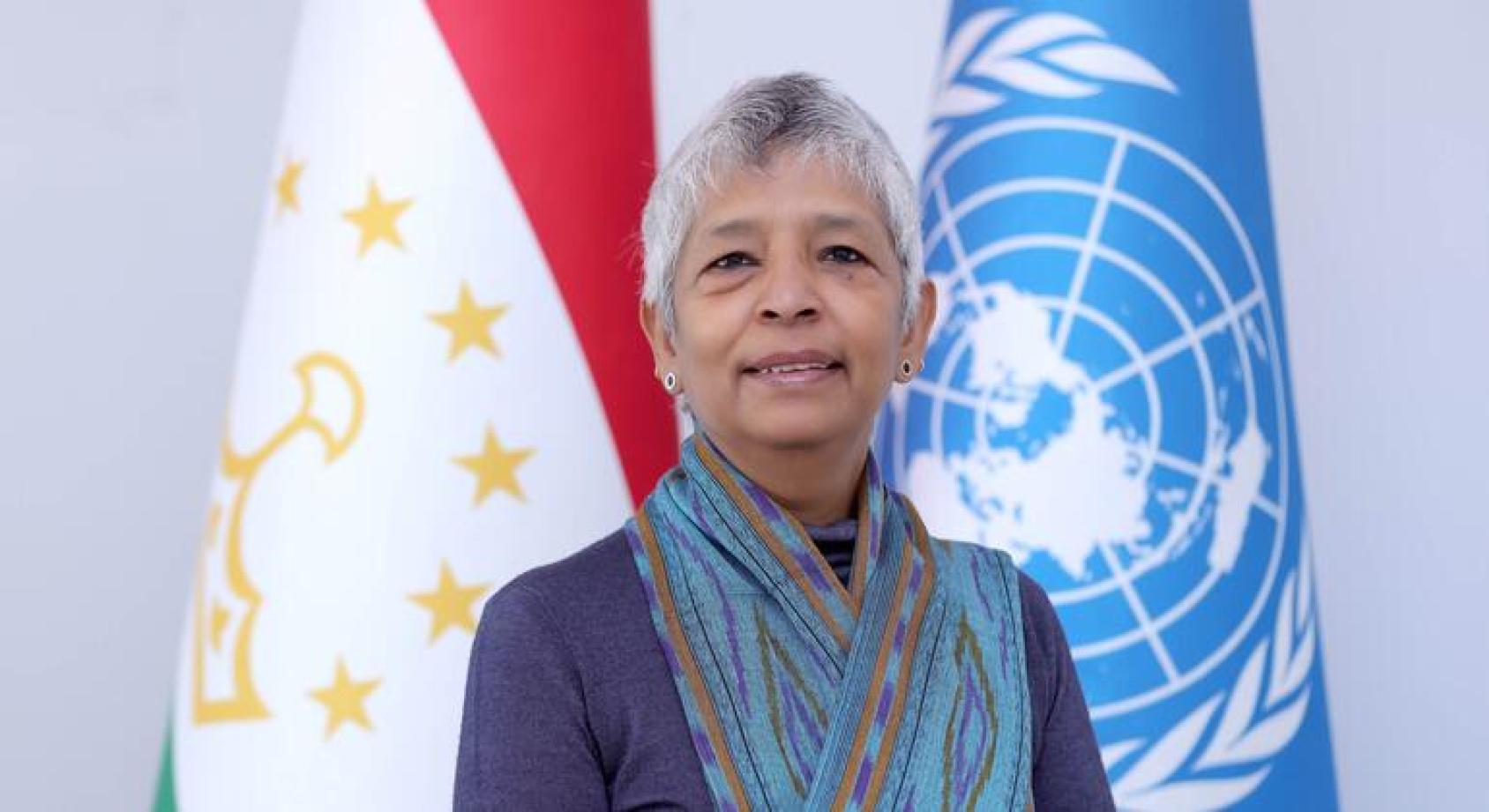 Портрет женщины с короткими седыми волосами, стоящей перед флагами Таджикистана и ООН.