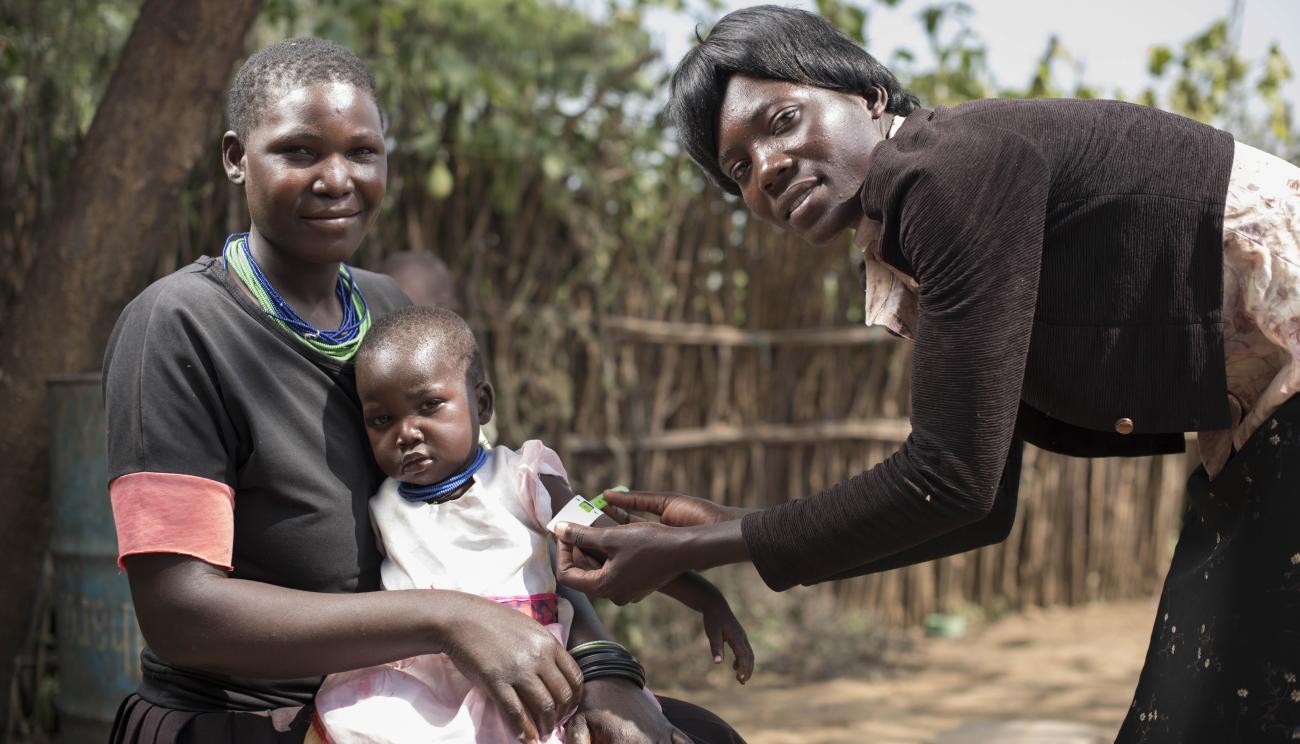 الأم تجلس مع ابنتها الصغيرة بينما تضع المرأة رباطًا حول ذراع الطفلة.