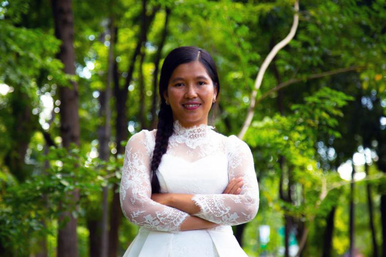 Shows Kanha standing with her arms crossed and an empowered smile facing the camera. She is wearing a white dress and stands in front of trees.