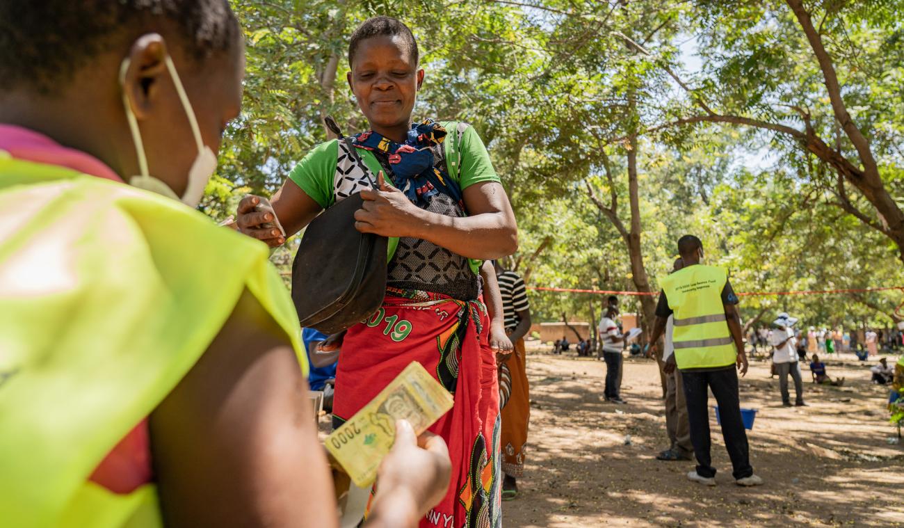 Une femme se tient debout en attendant de recevoir l’aide en espèces de l’ONU. Une femme est assise en face d’elle, dos à la caméra et compte des billets.