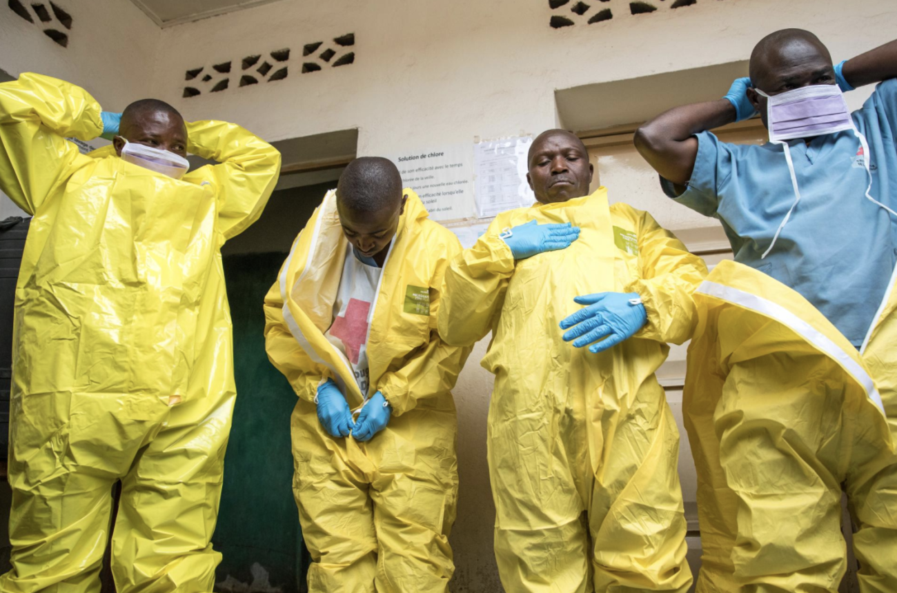 Quatre agents de santé se préparent en enfilant des combinaisons de protection jaunes, des masques chirurgicaux et des gants bleus.