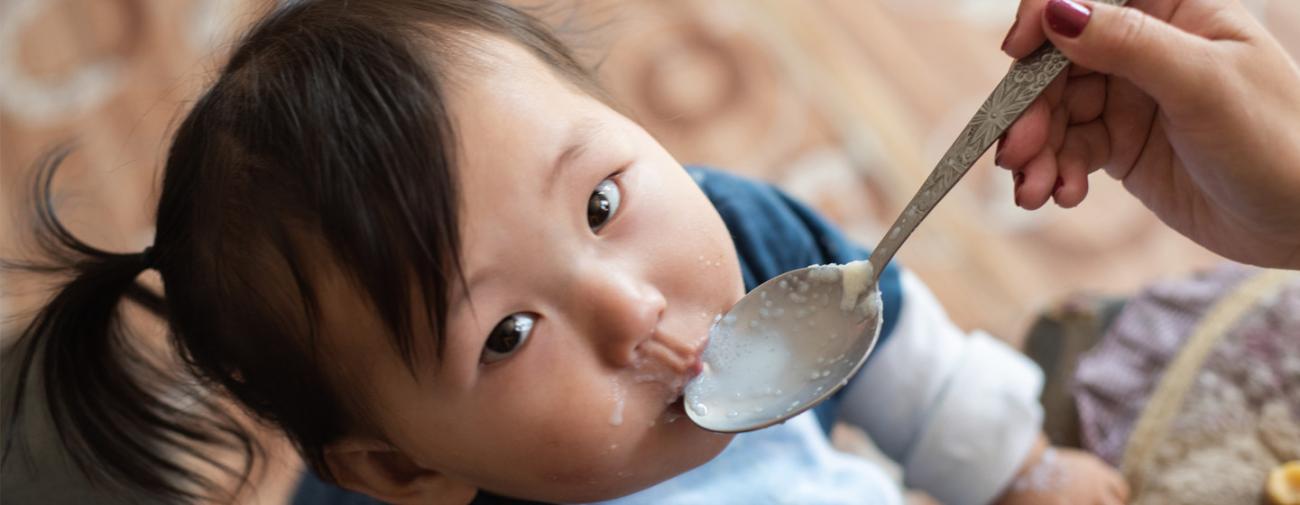 Gros plan sur un bébé qui regarde fixement la caméra alors qu'une personne hors-champ le nourrit à l'aide d'une cuillère.