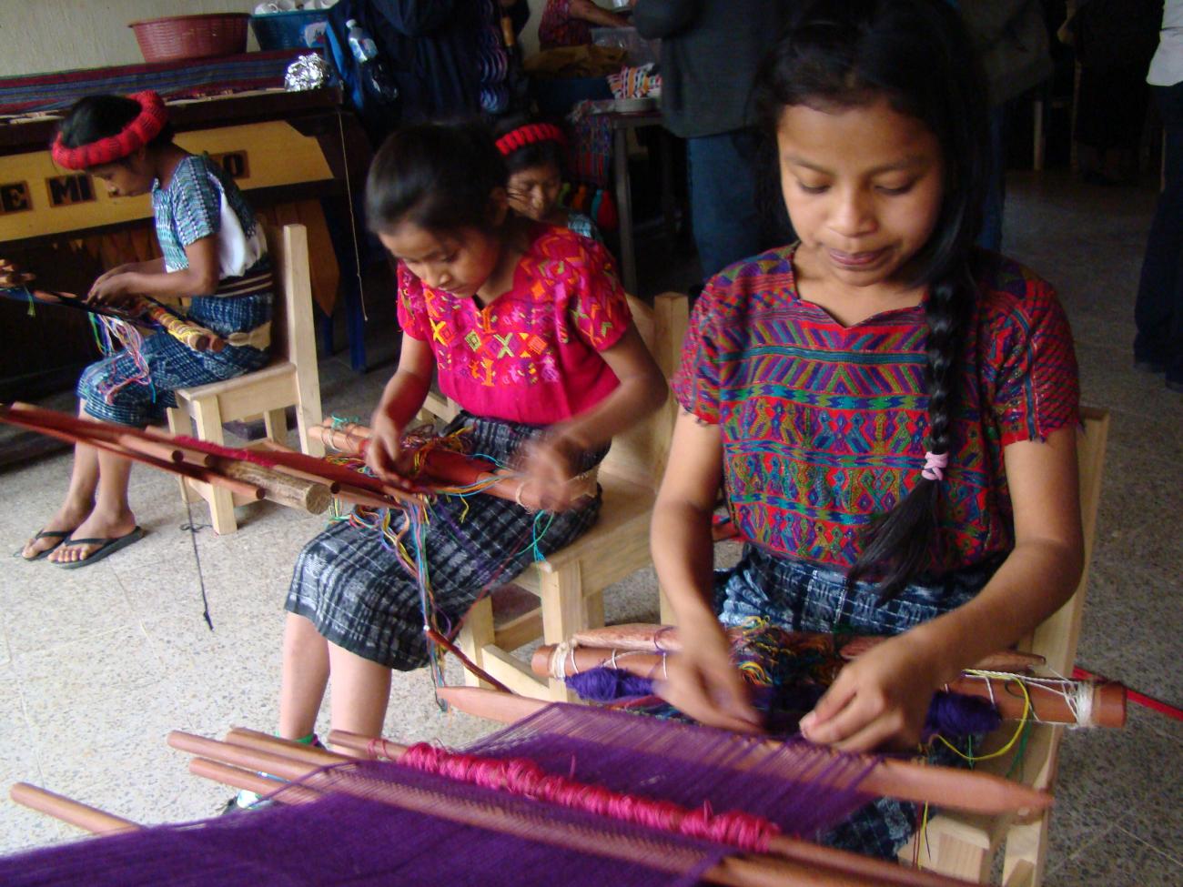 Photo showing three indigenous girls sitting on a chair and learning to weave using a traditional loom 