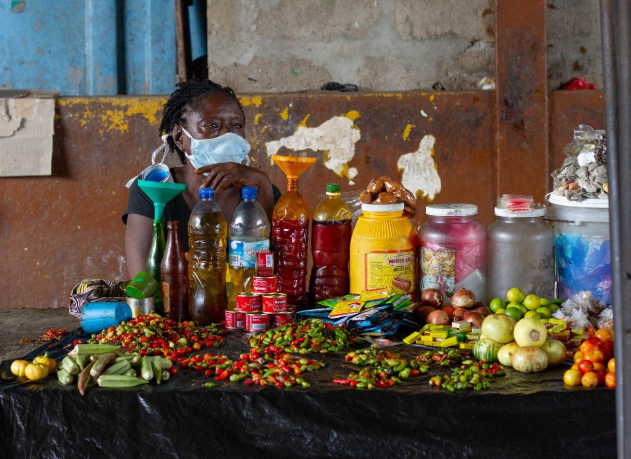 Une femme portant un masque de protection est assise derrière un étal du marché de Freetown. Sur son étal se trouvent toutes sortes de denrées alimentaires fraîches et en boîte.