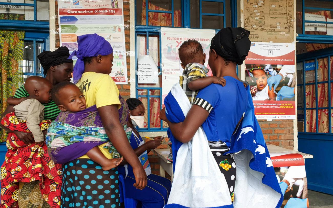Trois femmes portant chacune un bébé se tiennent devant la porte d'un centre médical et se renseignent auprès d’un membre féminin du personnel assis à une table à l’extérieur du centre. Plusieurs affiches contenant des informations sur la COVID-19 et sur la vaccination contre d’autres maladies infectieuses ont été collées sur le mur externe du centre.