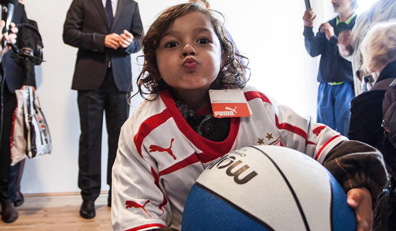 Un niño vestido con un chándal deportivo, sostiene una pelota de baloncesto con ambos brazos y mira a la cámara, haciendo el gesto de estar mandando un beso, con los labios fruncidos.
