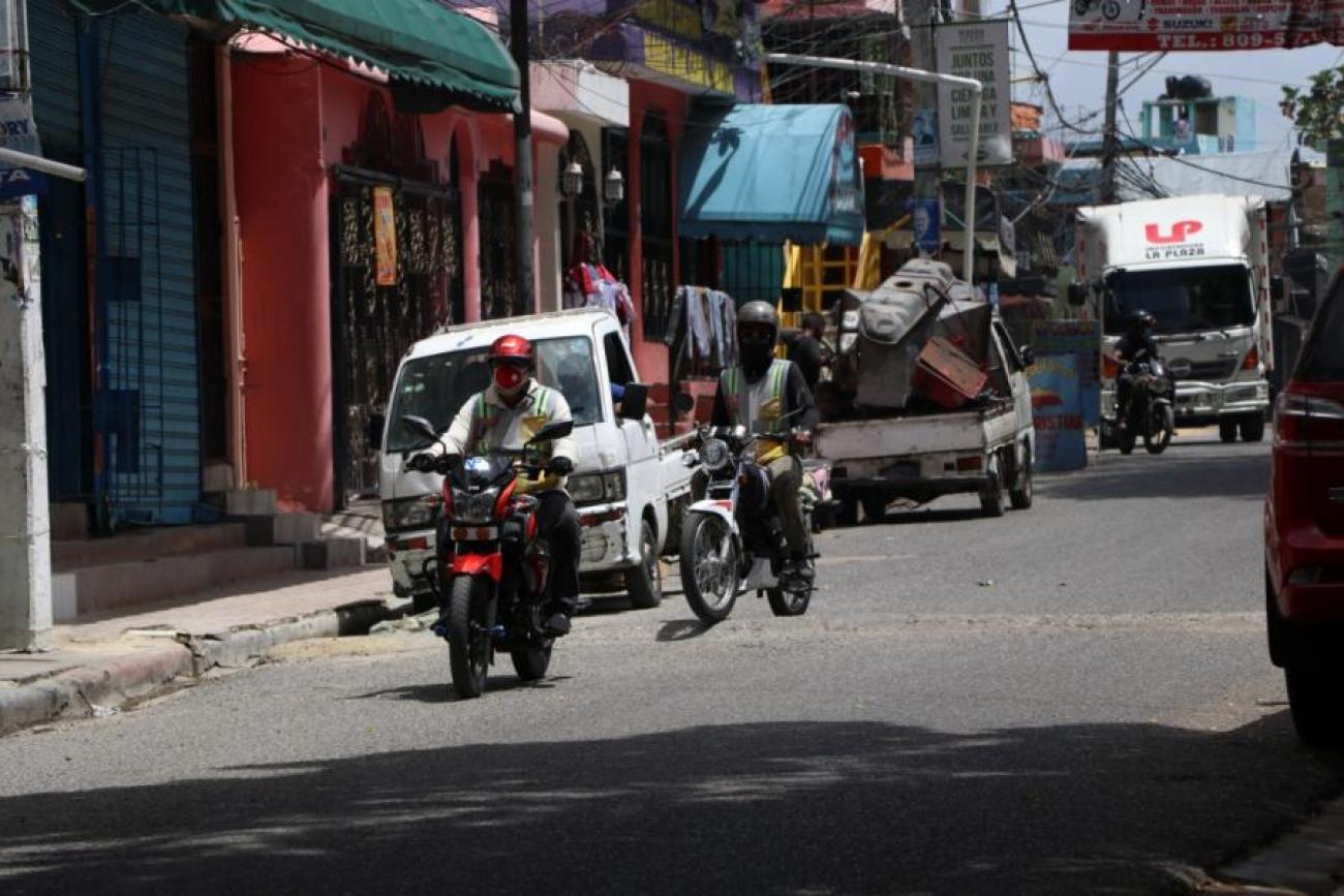 Escena de la vida citadina, donde se ve a motociclistas con protectores faciales y cascos de seguridad conduciendo por una calle; y varias camionetas estacionadas en los laterales de la calle.
