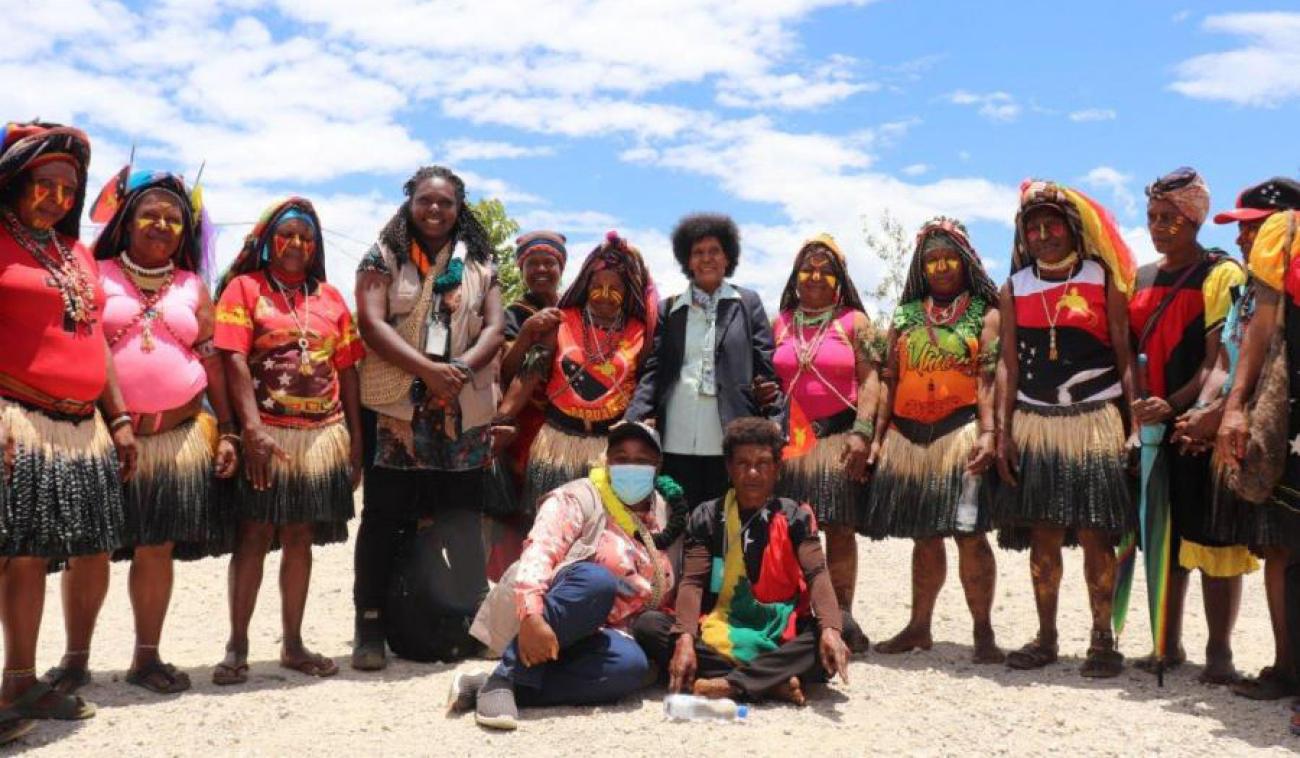 La delegación de ONU Mujeres y las mujeres miembros del Consejo Provincial de Hela están reunidas para la inauguración de la oficina de la ONU en Tari, provincia de Hela. La mayoría de las mujeres están de pie frente a la cámara, mientras que otras dos mujeres están sentadas en el suelo en primer plano. Muchas mujeres del consejo provincial visten ropas tradicionales de colores vivos.