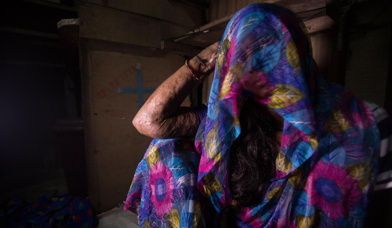 A woman sits on the floor and looks at the camera with the fabric from her dress over her head covering her face.