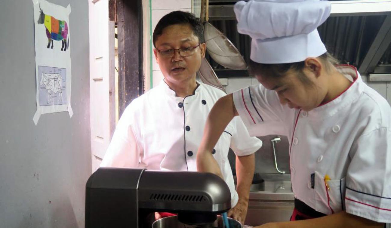 Una estudiante de cocina revuelve los ingredientes, manualmente. Al lado de ella, una persona con un traje de chef supervisa a la estudiante. En primer plano vemos la parte lateral de un electrodoméstico (ayudante de cocina, color negro).