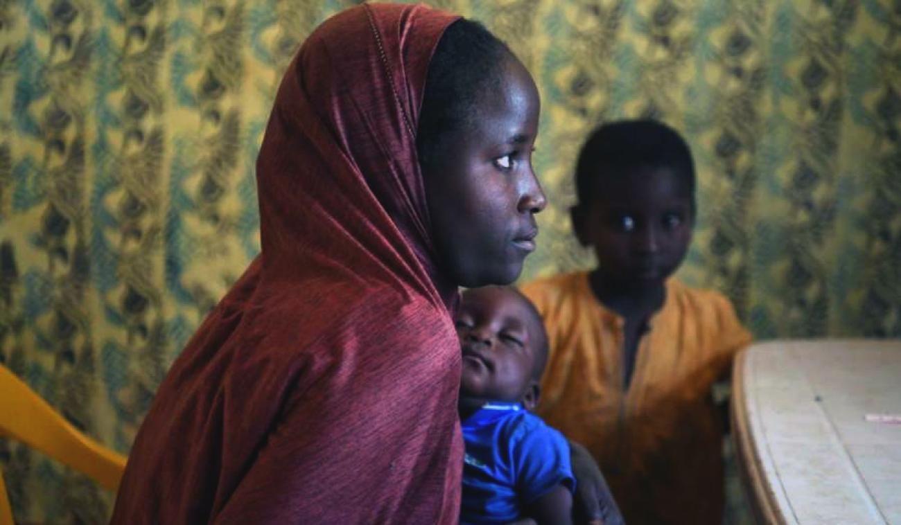 أم شابة تحمل طفلها النائم، بينما يظهر فتى يلبس قميصًا أصفر في الخلفية.