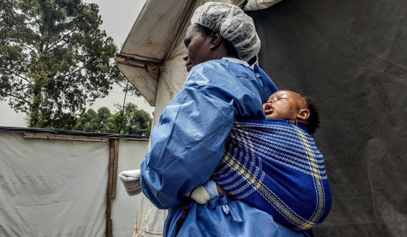 امرأة ترتدي معدات الوقاية الشخصية وتحمل طفلاً صغيراً على ظهرها.