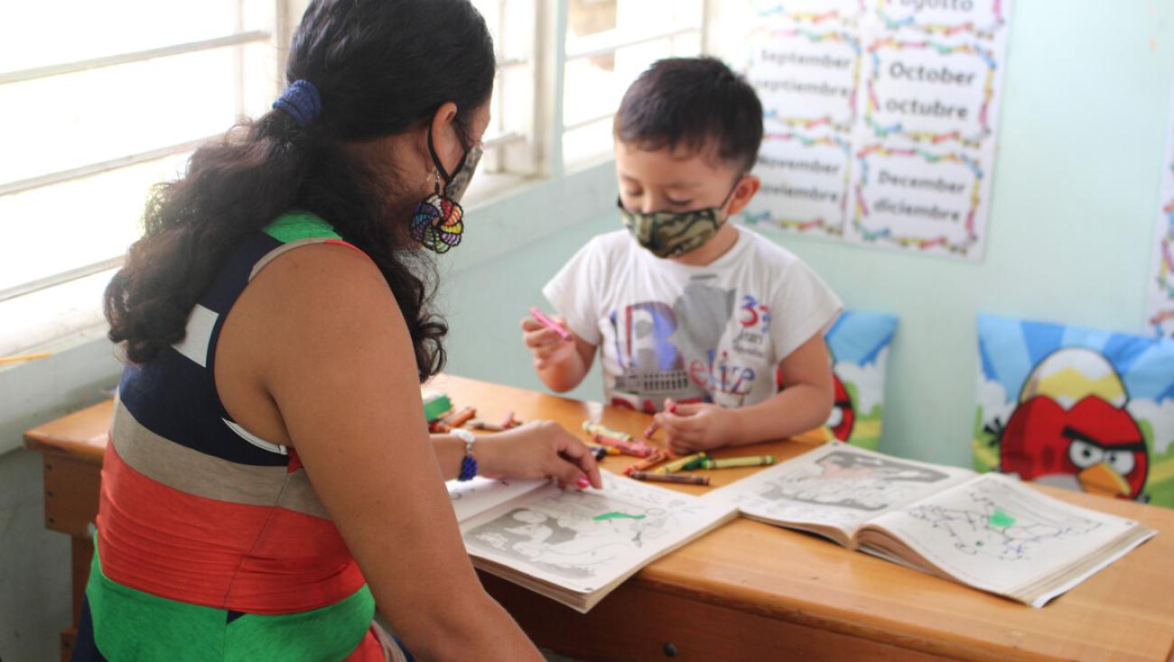 Une femme et un enfant portant des masques de protection sont assis à une table en bois l’un en face de l’autre et regardent des livres de coloriage.