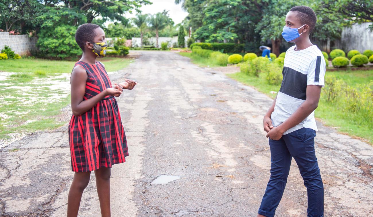 Una niña y un niño adolescentes con mascarillas demuestran su distanciamiento social al separarse mientras conversan, en medio de una carretera de tierra en una zona verde, en Lusaka, Zambia.