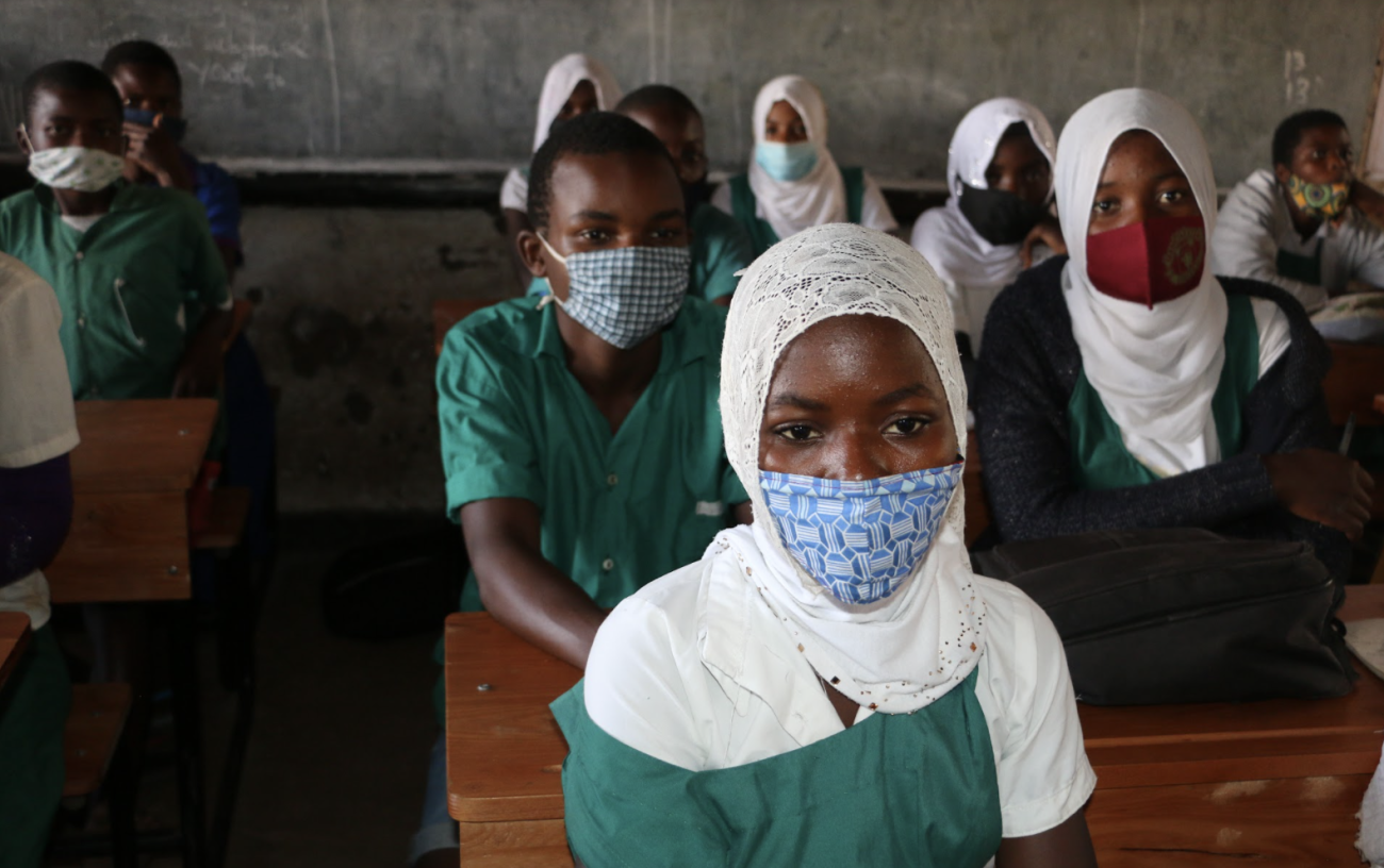 مجموعة من الأطفال يرتدون الزي المدرسي والكمامات يجلسون في فصل دراسي وينظرون إلى الأمام.