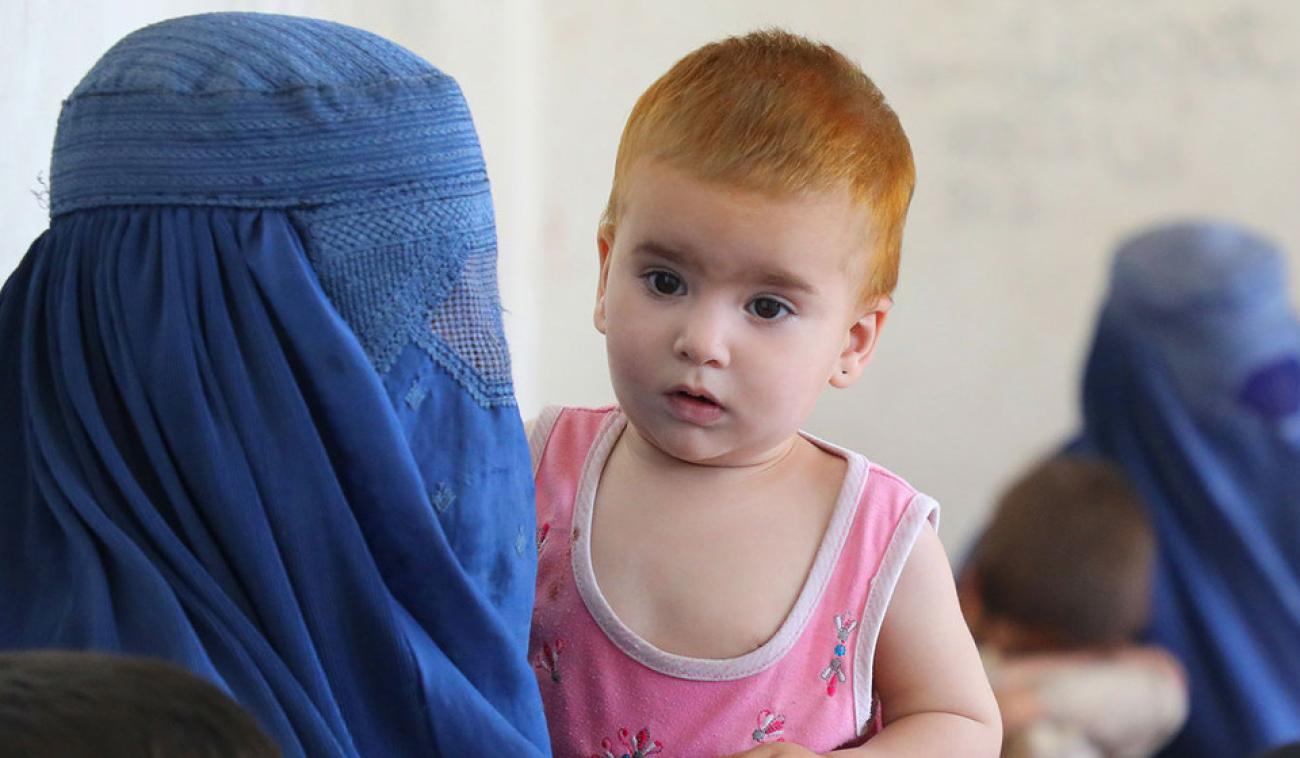 En primer plano aparece una madre con tocado y rostro mientras sostiene a su bebé, y en segundo plano se encuentra otra madre y su pequeño.